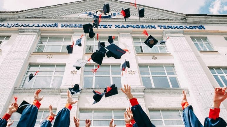 school graduates standing in front of school throwing their graduation caps in the air